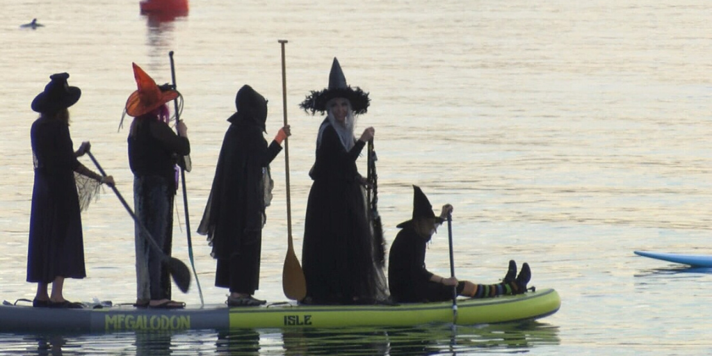 Four backlit witches stand on SUP with paddles. One warlock sits on the front of the paddle board to steer